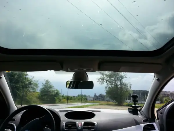 Widok przez panoramiczny dach samochodu. Krople deszczu na szybie, droga i drzewa w tle.