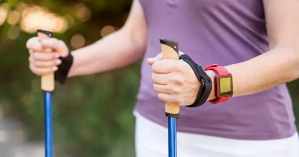 Dłonie trzymające kijki do nordic walking, z czerwonym zegarkiem na nadgarstku. To świetny sposób, jak chodzić z kijkami, by wzmocnić ciało.