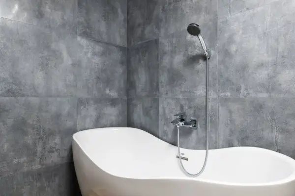 Nowa, wolnostojąca wanna o ergonomicznym kształcie, z baterią i słuchawką prysznicową, na tle nowoczesnych, szarych płytek. Szerokość wanny pozwala na komfortową kąpiel.