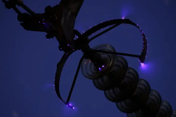 Des arcs électriques violets, les feux de Saint-Elme, crépitent sur une structure métallique et des isolateurs en verre contre un ciel nocturne.