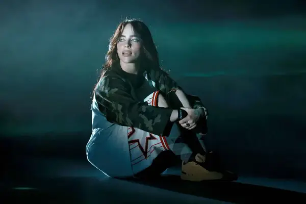 Billie Eilish siedzi na scenie, wpatrzona w dal. Jej stylizacja i mroczny klimat sugerują, że zaraz usłyszymy fajne piosenki.