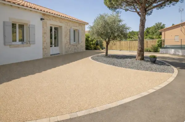 Allée de maison avec béton désactivé de belle épaisseur, bordée de pierres et de gravier sombre autour d'un arbre.