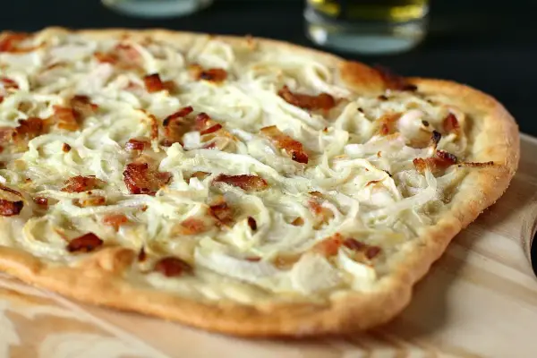 Flammekueche généreuse garnie d'oignons crus ou cuits et de lardons croustillants, prête à être dégustée.