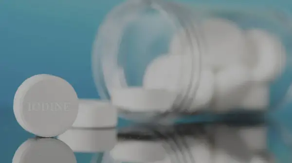 Pastillas de yodo blancas, una en primer plano con la palabra "IODINE" grabada, y un frasco volcado con más pastillas.