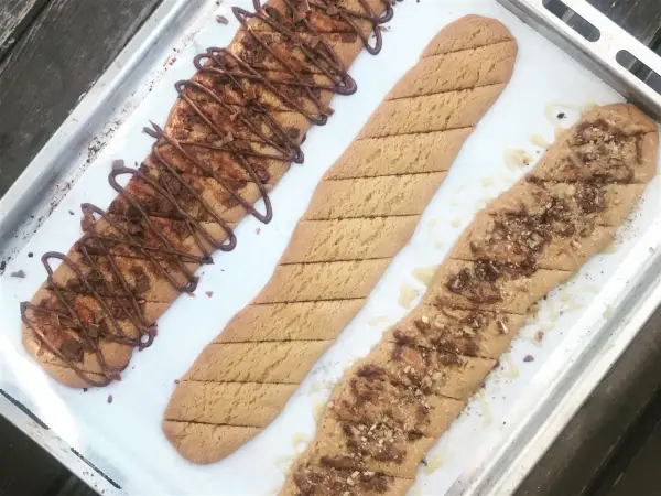 Tre långa, avlånga kakor på en plåt. En är dekorerad med chokladglasyr och hackad choklad, en annan med strössel och en tredje med honung och nötter. Perfekt för ett kolakakor recept!