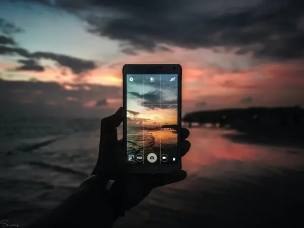 Mano che tiene un cellulare per scattare una foto con tramonto colorato sul mare.