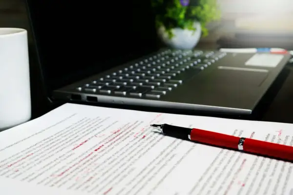 Czerwony długopis poprawia tekst na papierze, obok laptopa. Widać cudzysłów odmiana i poprawki.