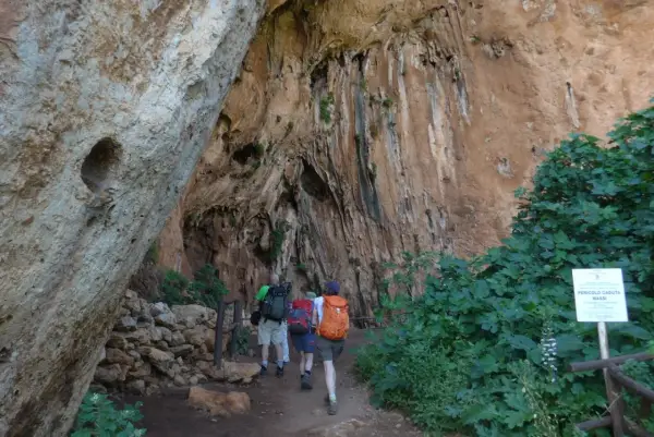 Escursionisti esplorano un sentiero nella Riserva dello Zingaro, avvicinandosi a un'imponente parete rocciosa con vegetazione rigogliosa.