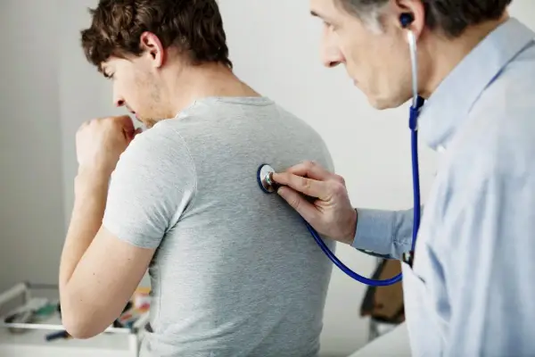 Arzt untersucht Patienten mit Stethoskop. Der Patient hustet, was auf Kurzatmigkeit hindeuten könnte.