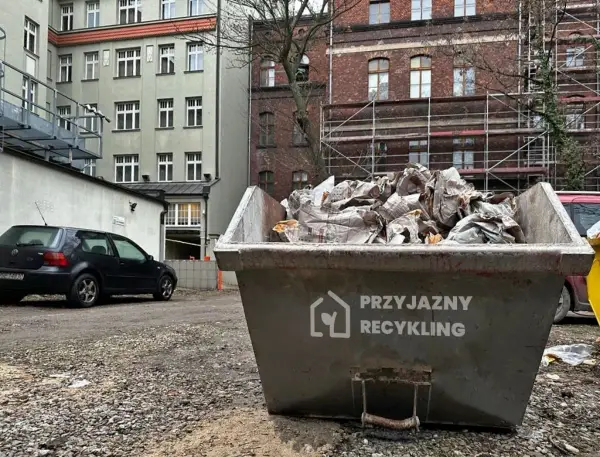 Kontener na odpady budowlane, kod odpadu 17 09 04. Wypełniony gruzem i opakowaniami, obok stoi czarny samochód.