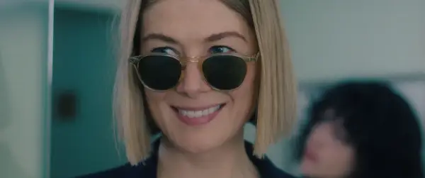 Uśmiechnięta Rosamund Pike w okularach przeciwsłonecznych, przypominająca scenę z jej filmów i seriali.