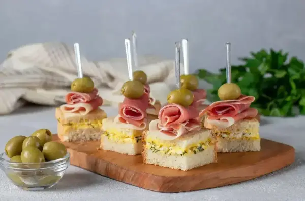 Eleganckie mini przekąski na imprezę: kanapki z szynką, jajkiem i oliwkami na wykałaczkach.