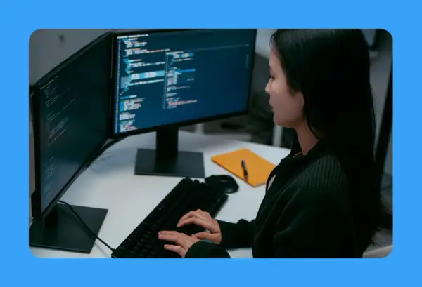 Jeune femme travaillant sur un ordinateur, avec deux écrans affichant du code. Sa position devant l'écran du bureau est concentrée.