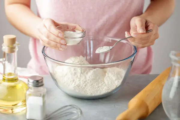 Une personne ajoute une poudre blanche à de la farine dans un bol. Une cuillère et un petit bol sont utilisés. C'est une idée pour par quoi remplacer la levure chimique.