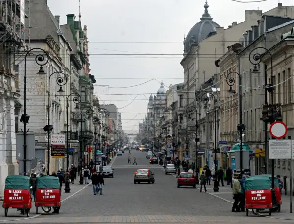 Ulica Piotrkowska w Łodzi, pełna zabytkowych kamienic, latarni i ruchu ulicznego. Dwa czerwone pojazdy z napisem "Miejsce na reklamę" stoją przy chodniku.
