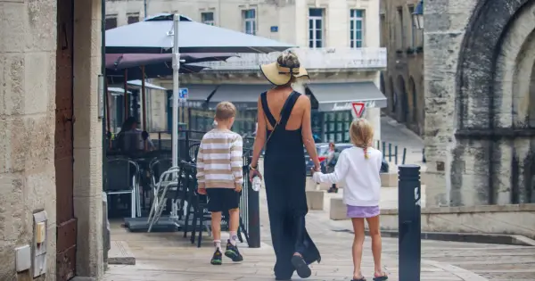 Kobieta w letniej sukience i kapeluszu prowadzi dwójkę dzieci, chłopca i dziewczynkę, ulicą. To francuskie dziecko, które zwiedza miasto.