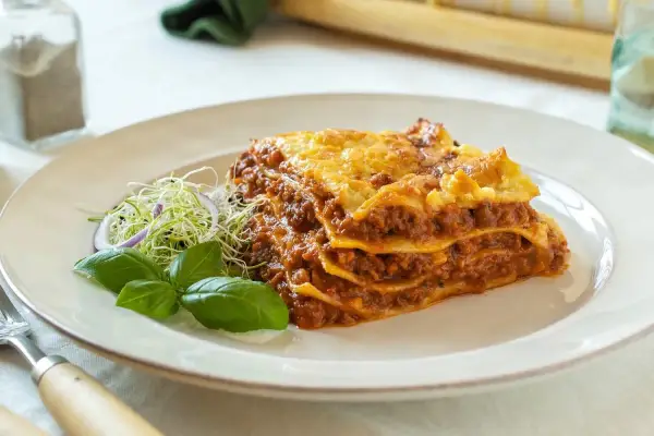 Porción de lasaña de carne casera, con capas de pasta, salsa boloñesa y queso gratinado, servida con brotes y albahaca.