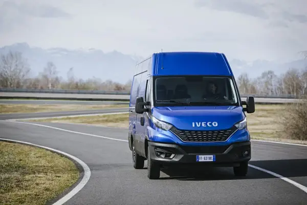 Niebieski Iveco Daily 3 na zakręcie drogi, z kierowcą za kierownicą. W tle góry i drzewa.