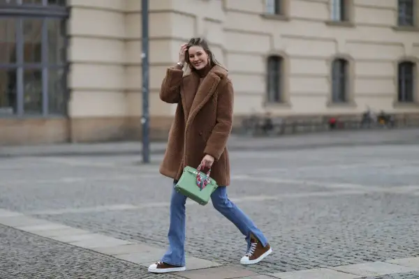 Frau in braunem Teddy-Mantel, blauer Jeans und braunen Schuhen mit weißer Sohle geht lässig über Kopfsteinpflaster.