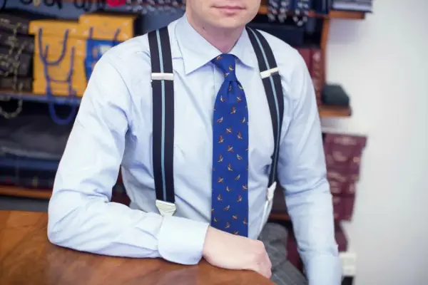 Elegancki mężczyzna w błękitnej koszuli i krawacie z ptakami, pokazuje, jak nosić szelki.