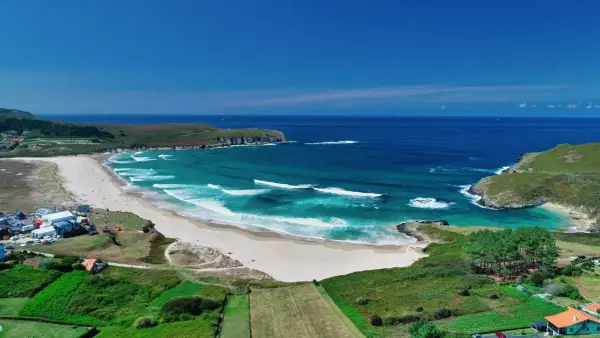 Vista aérea de la playa de Pantín con olas turquesas rompiendo en la arena dorada, rodeada de verdes colinas y el azul intenso del océano.