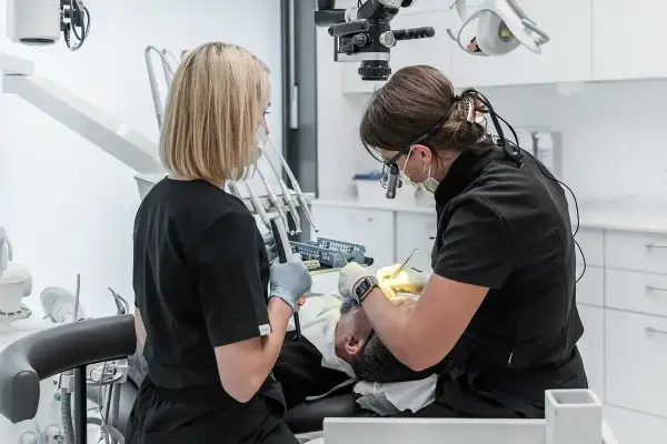 Dwie dentystki pracują przy pacjencie. Jedna używa mikroskopu, druga trzyma narzędzie. To obrazek pokazujący, jak zostać dentystą.