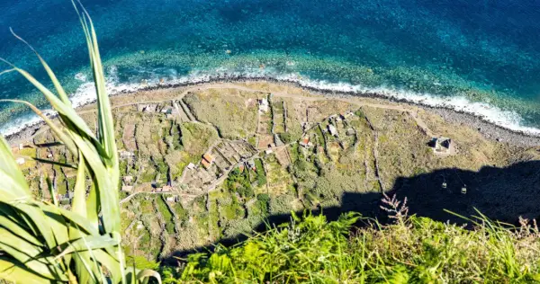 Terraced fields and houses dot the coastline, a glimpse of what to see in Madeira. The turquoise ocean crashes against the rocky shore.