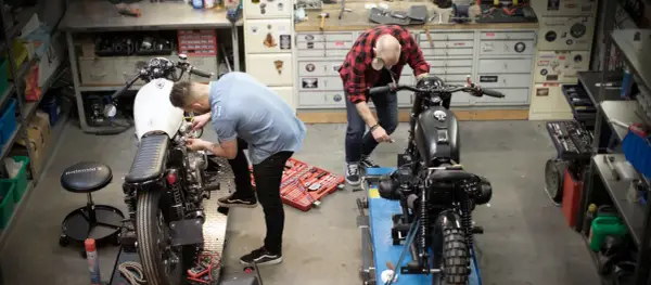 Due uomini lavorano su due moto in un garage. Uno è chino su una moto bianca, l'altro su una nera. Un kit di attrezzi è aperto sul pavimento. Progetto schienalino moto fai da te.