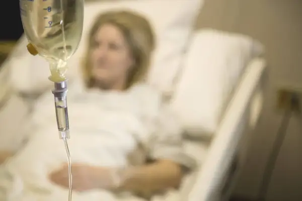 Infusion vorbereiten: Eine Frau liegt im Krankenhausbett, während eine Infusionstropfflasche und ein Tropfschlauch im Vordergrund zu sehen sind.