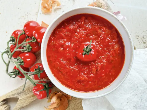 Eine Schüssel mit köstlicher, hausgemachter italienischer Tomatensauce, garniert mit einer frischen Kirschtomate. Daneben liegen frische Tomaten und Zwiebeln.