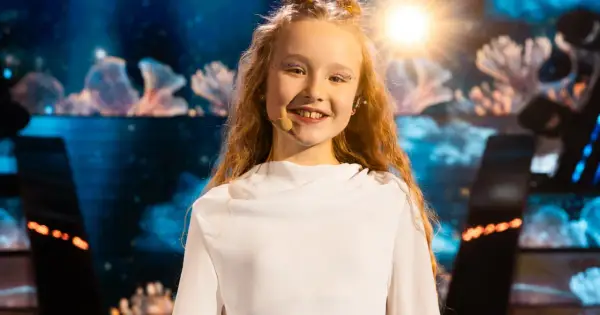Uśmiechnięta dziewczynka z błyszczącym makijażem na oczach, w białej sukience, gotowa do występu na Eurowizji Junior Polska.