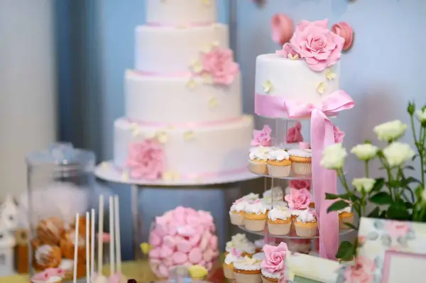 Eleganckie, białe torty weselne i babeczki ozdobione różowymi kwiatami i wstążkami. Idealne na słodki stół, inspiracja jakie ciasta na wesele.
