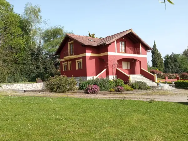 Una casa típica cántabra, de color rojo intenso con detalles amarillos, rodeada de vegetación y un césped verde.