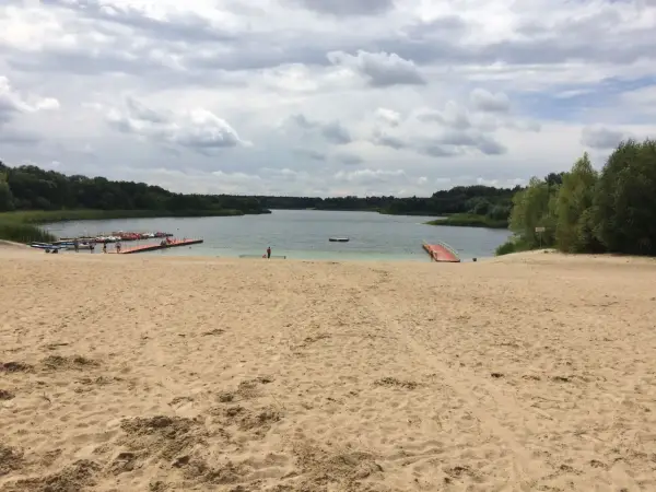 Piaskowa plaża nad jeziorem, z pomostami i łódkami. W oddali las, chmury na niebie. Idealne miejsce na wypoczynek nad wodą.