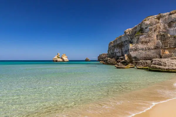 Spiaggia dorata e mare cristallino in Puglia, con faraglioni che emergono dalle acque turchesi e scogliere imponenti.