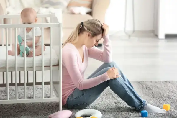 Zmęczona mama siedzi na podłodze obok łóżeczka, czując baby blues. Dziecko bawi się zabawką.