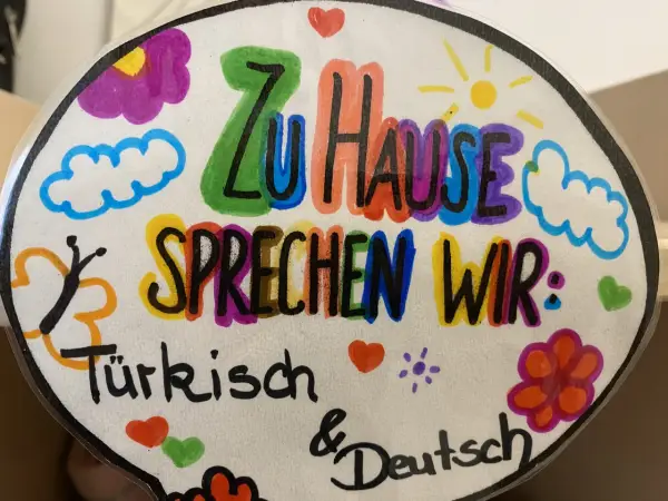 Bunte Zeichnung mit der Aufschrift "Zu Hause sprechen wir: Türkisch & Deutsch". Ein Beispiel für vorurteilsbewusste Pädagogik, die Mehrsprachigkeit feiert.