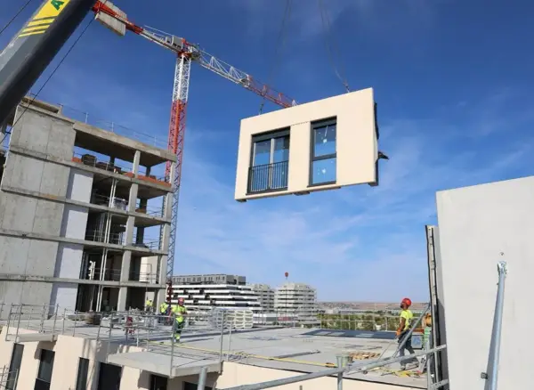 Grúa elevando un módulo prefabricado para la construcción industrializada de un edificio. Obreros supervisan la operación.