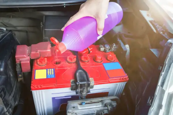 Ręka wlewa fioletowy płyn, prawdopodobnie elektrolit do akumulatora, do czerwonej obudowy akumulatora samochodowego.