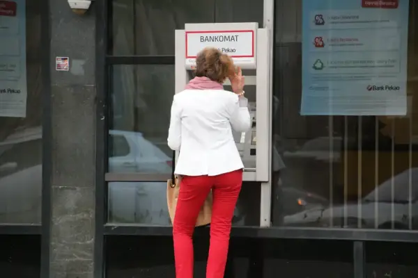 Kobieta w czerwonych spodniach i białej marynarce korzysta z bankomatu. Może to być symboliczne przedstawienie średnich zarobków w Polsce, gdzie ludzie polegają na bankomatach.