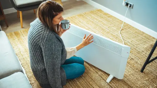 Mujer con taza de té cerca de un calefactor eléctrico, una solución para la alternativa al gas en un piso.