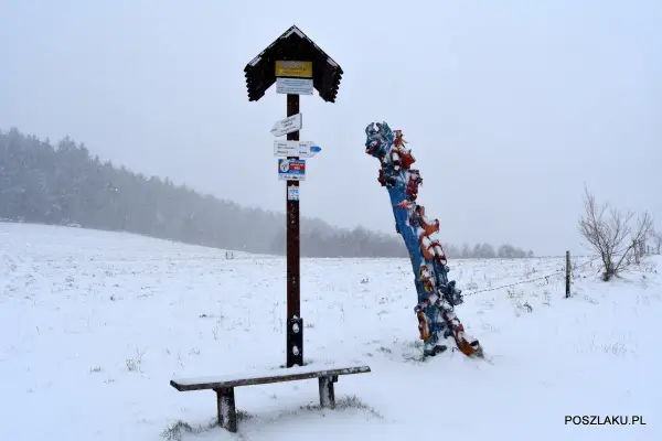 Zimowy krajobraz z ławką i drogowskazem turystycznym. Obok kolorowa rzeźba, być może symbolizująca skopiec szczyt.