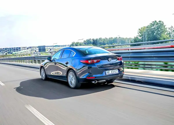 Ciemnoniebieska Mazda 3 sedan jedzie po autostradzie, odbijając światła i otoczenie.