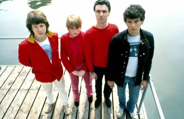 Die Band Talking Heads steht auf einem Holzsteg. David Byrne trägt ein rotes Hemd, Tina Weymouth ein pinkes Outfit.