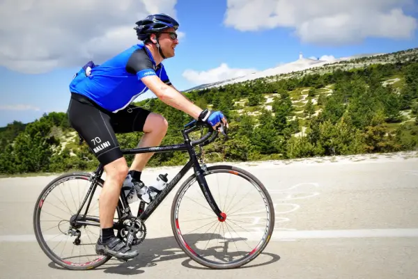Ciclista in maglia blu affronta la **salita** del **Mont Ventoux**, con la cima innevata in lontananza.