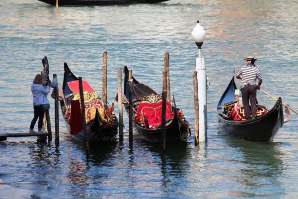 Gondole veneziane ornate, una turista scatta foto. La differenza tra gondola e sandalo è chiara: una naviga, l'altro si indossa.