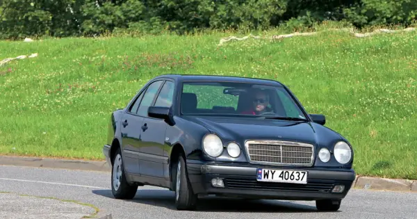 Czarny Mercedes W210 z silnikiem om606 jedzie po drodze, za nim zielone, kwieciste wzgórze.