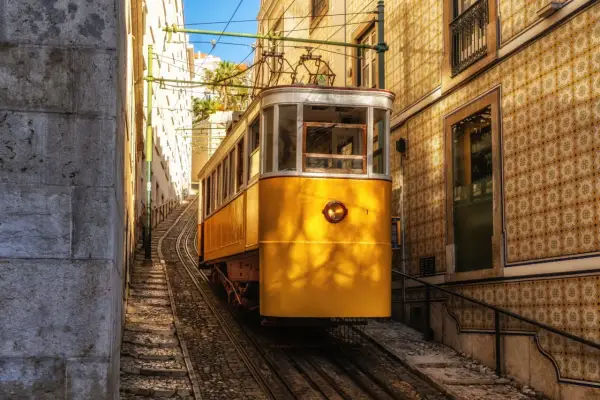 Żółty tramwaj wspina się po stromych torach w wąskiej uliczce Lizbony. W tle widać kamienne budynki pokryte tradycyjnymi płytkami azulejo.
