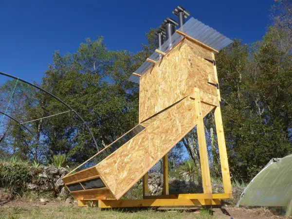 Un sechoir solaire artisanal en bois, avec un panneau incliné vitré, sous un ciel bleu.