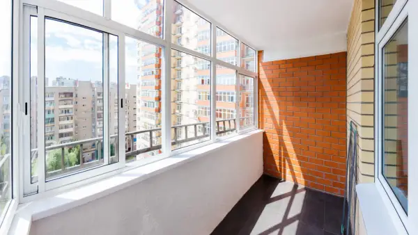 Wnętrze loggii z widokiem na miejską zabudowę. Ściany z cegły i tynku, przeszklony balkon z białymi ramami okiennymi.